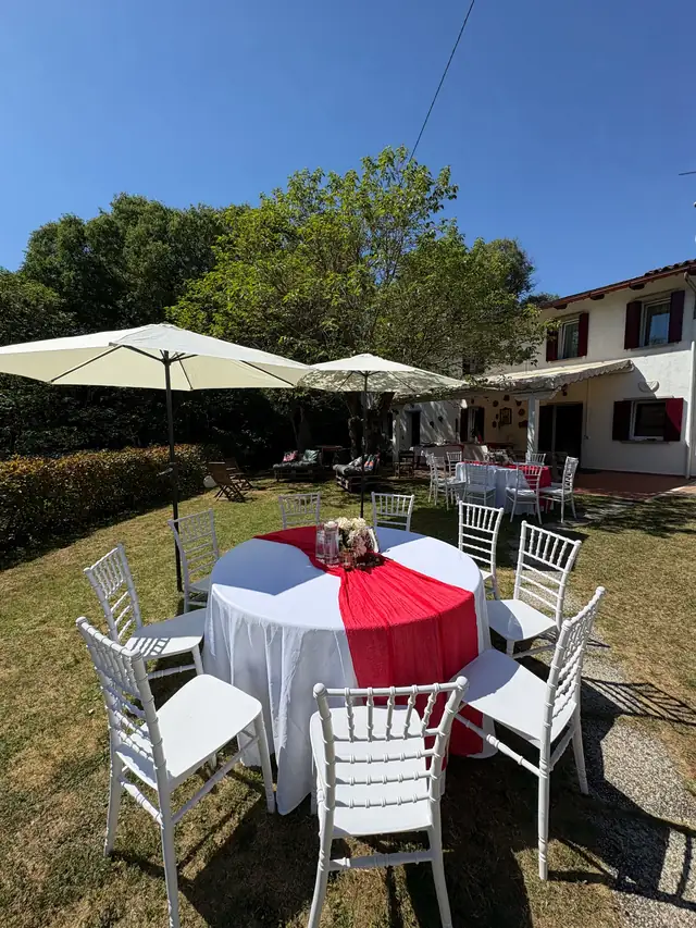 Tavolo rotondo con drappo rosso e sedie bianche in giardino, con ombrelloni e villa sullo sfondo